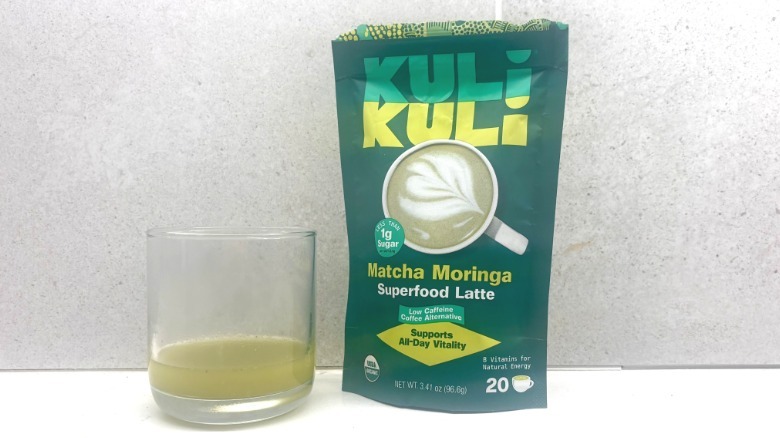 A glass of green matcha next to a sealed bright green bag of Kuli Kuli matcha powder on a countertop.