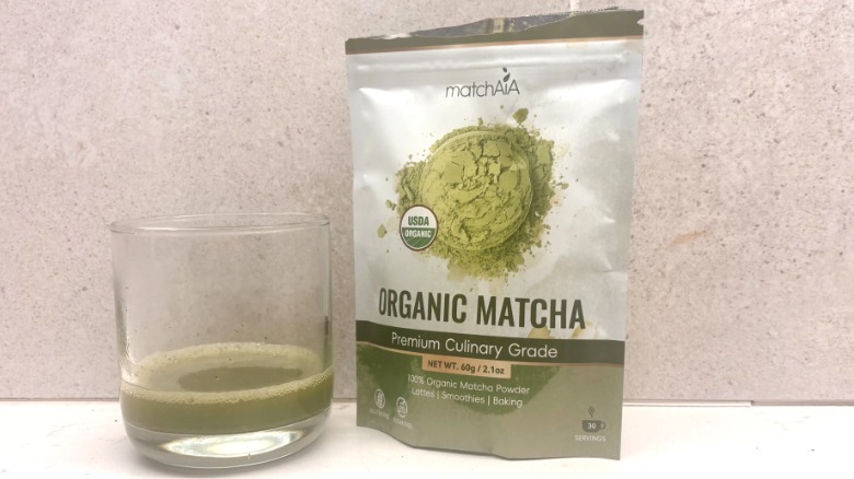 A glass filled with green matcha sits beside a sealed white and green bag of matchAIA matcha powder on a countertop.
