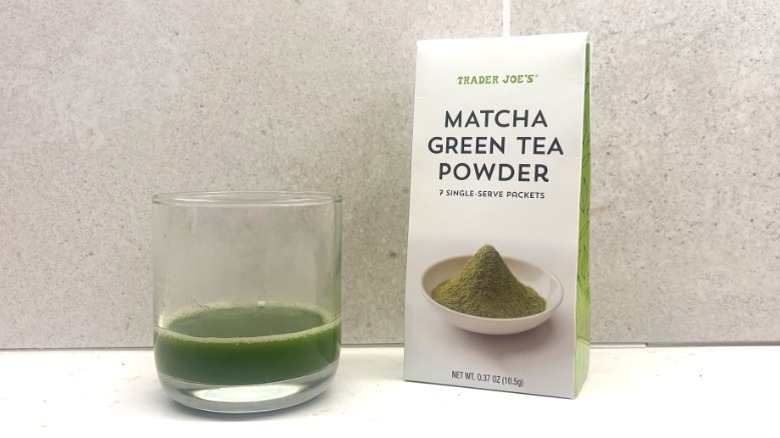 A glass of green matcha beside a white box of Trader Joe's matcha powder featuring an image of matcha on the front on a countertop.