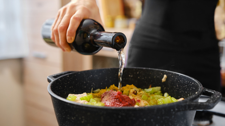 A person pouring wine into a pot with sauce and vegetables