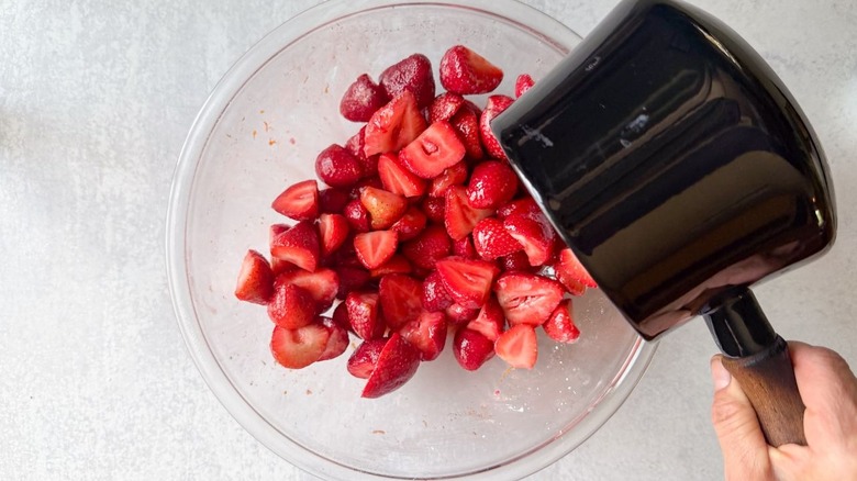 Pouring hot berry syrup over strawberry halves in glass bowl