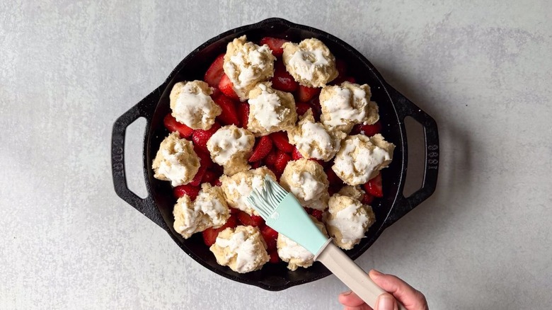 Brushing heavy cream on biscuit dough over strawberry cobbler filling in cast iron skillet