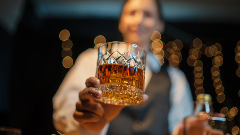 A glass of whiskey being held by an out-of-focus bartender