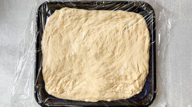 Plastic-covered focaccia dough rising in pan on countertop