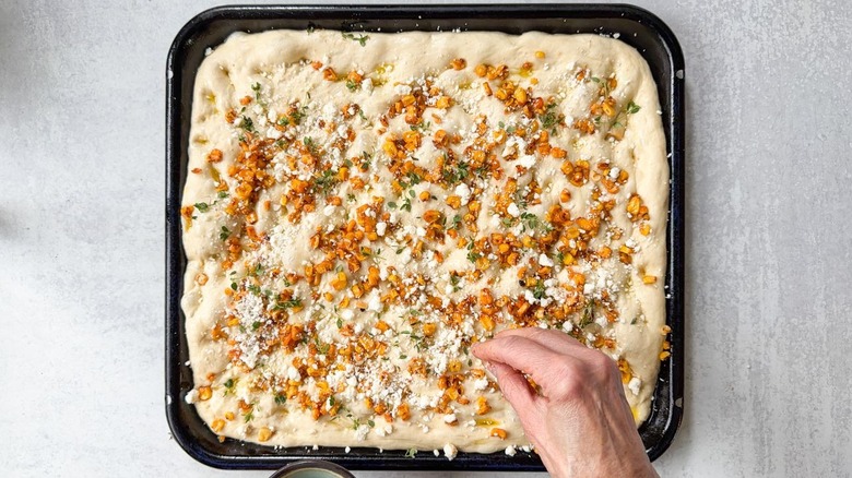Adding roasted corn and cotija cheese to dimpled focaccia dough on blue baking sheet