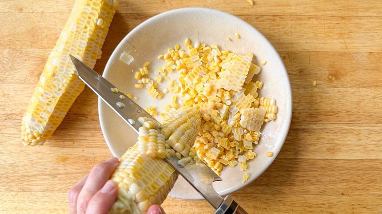 Cutting corn kernels away from cobs into shallow bowl on cutting board