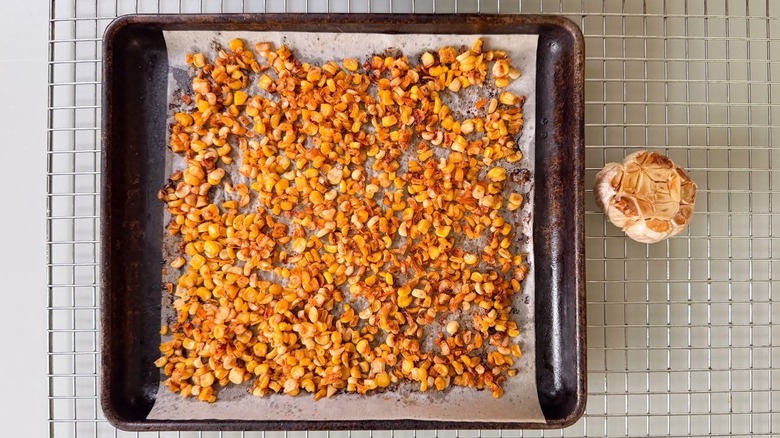 Roasted seasoned corn kernels on parchment-lined baking sheet and a head of roasted garlic on cooling rack