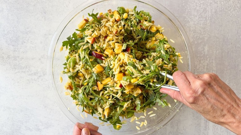 Tossing summery grilled corn and orzo salad with dressing using tongs in glass bowl