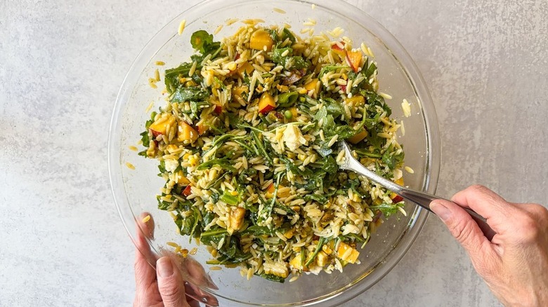 Folding crumbled cotija cheese into summery grilled corn and orzo salad using fork in glass bowl