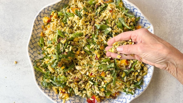 Sprinkling crushed pistachios on summery grilled corn and orzo salad in serving dish