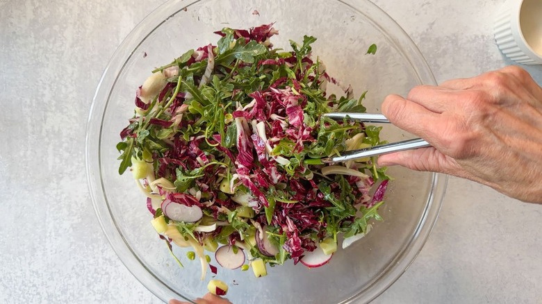 Tossing salad greens and vegetables with dressing in glass bowl with tongs