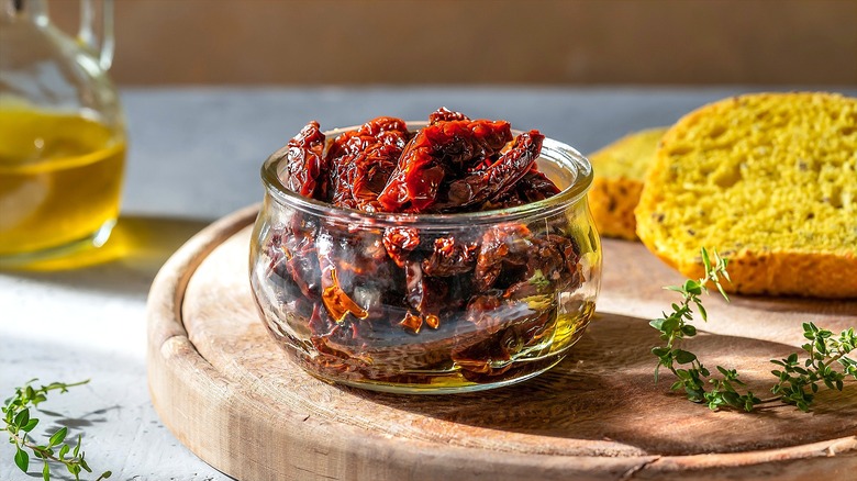 Small clear jar filled with sun-dried tomatoes in oil on a wooden serving board with sliced bread