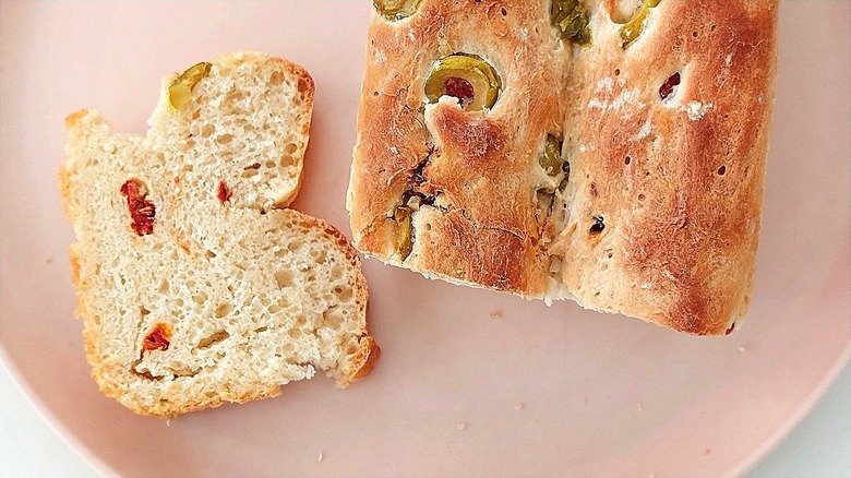Loaf and slice of quick bread with green olives and sun-dried tomatoes on a pastel pink plate