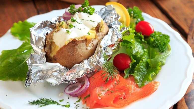 Split baked potato with sour cream and chives in foil on a white plate with smoked salmon, red onion, lettuce, and cherry tomatoes