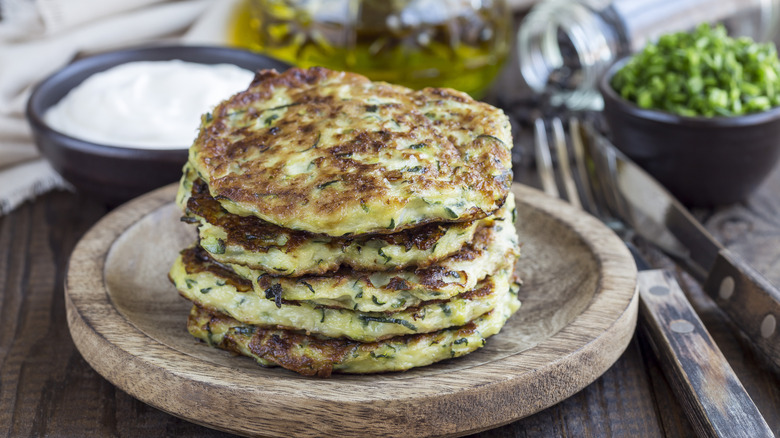 Vegetarian zucchini fritters or pancakes stacked on a wooden plate with various toppings behind