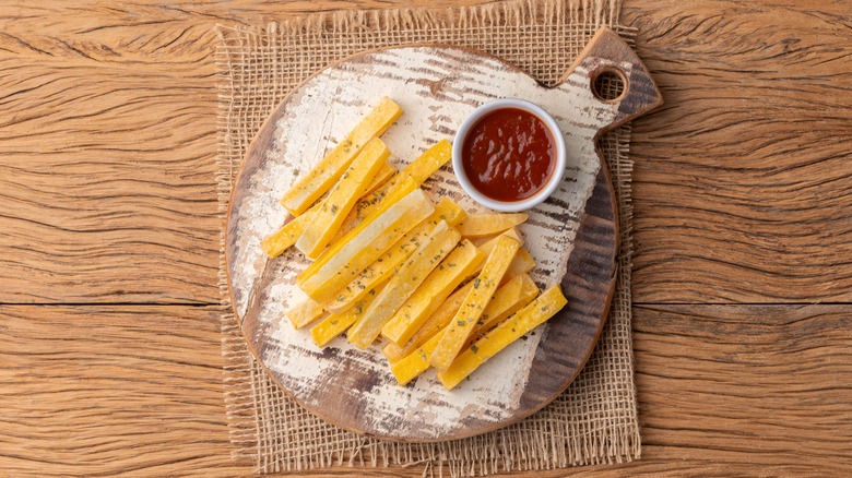 Crispy polenta fries and a bowl of ketchup on a wooden chopping board with wooden table background