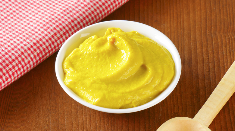 A small bowl of yellow mustard against a wood background with a folded picnic blanket.