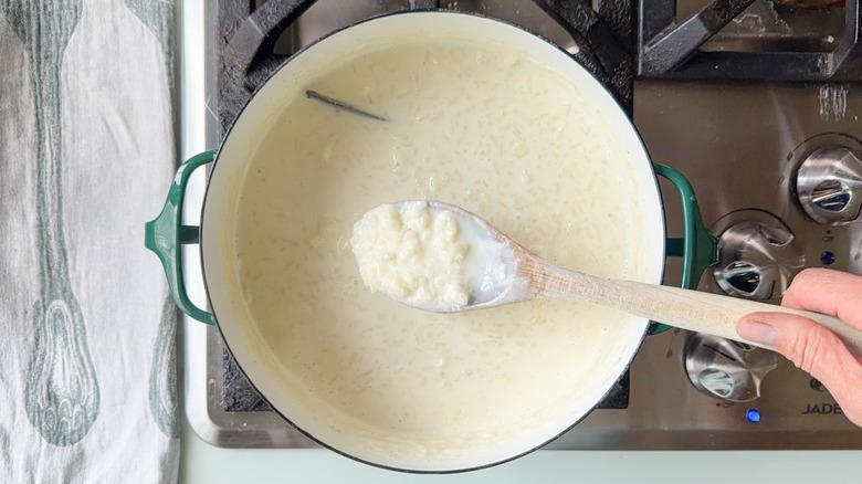 Wooden spoon filled with cooked arborio rice in milk with vanilla bean in large saucepan on stovetop