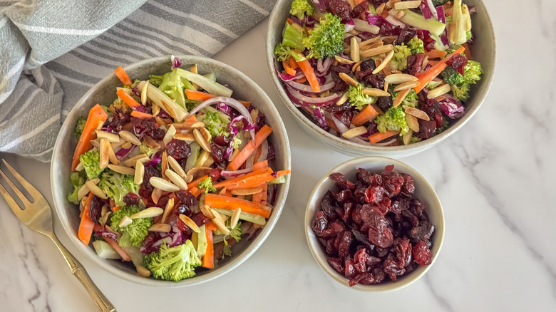 Bowls of broccoli slaw next to bowl of dried cranberries
