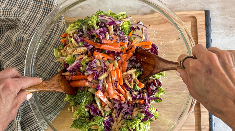 Hands using wooden spoons to toss broccoli slaw together in bowl
