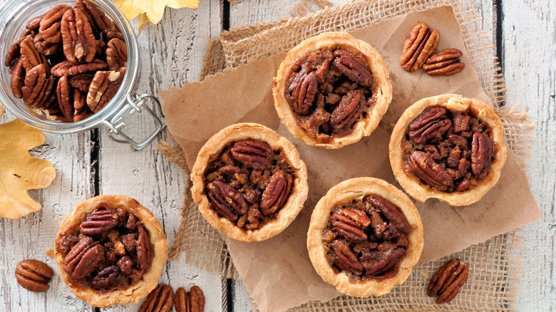 Mini pecan pies beside a glass jar of pecans on a wooden table.