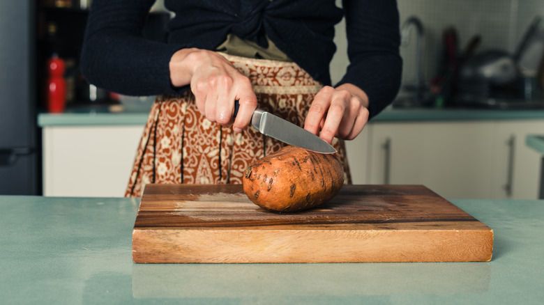 A person's hands are seen cutting a sweet potato in half with a sharp knife on a wooden cutting board