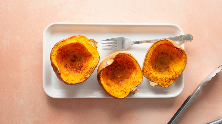 Three halves of roasted honeynut squash on a rounded rectangular tray with a stainless steel fork and some metal tongs off to the side