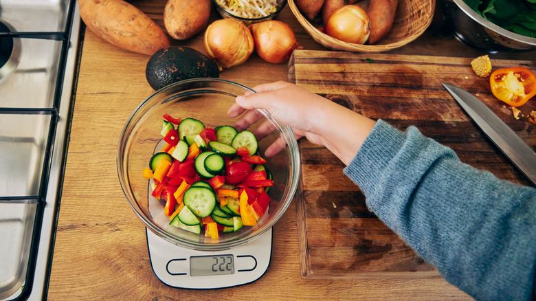 Person holding glass bowl filled with sliced vegetables on a food scale with other ingredients for cooking nearby