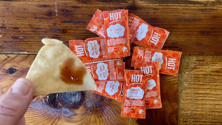 Hand holding tortilla chip dipped in Taco Bell Hot Sauce with packets in the background