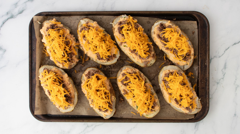 Potato skins on a large baking sheet filled with seasoned mashed potato, taco beef and grated cheddar