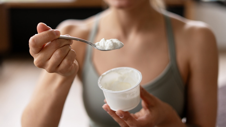 Close-up of a woman dressed in green sports clothing holding a spoon with fresh yogurt in one hand, and in the other hand carrying a yogurt cup