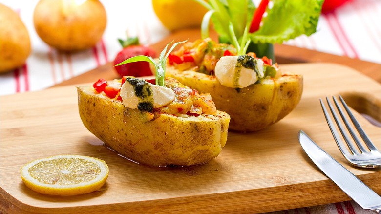 Two baked potato halves topped with sour cream, sauteed veggies, green sauce, and lemon juice on a wooden cutting board