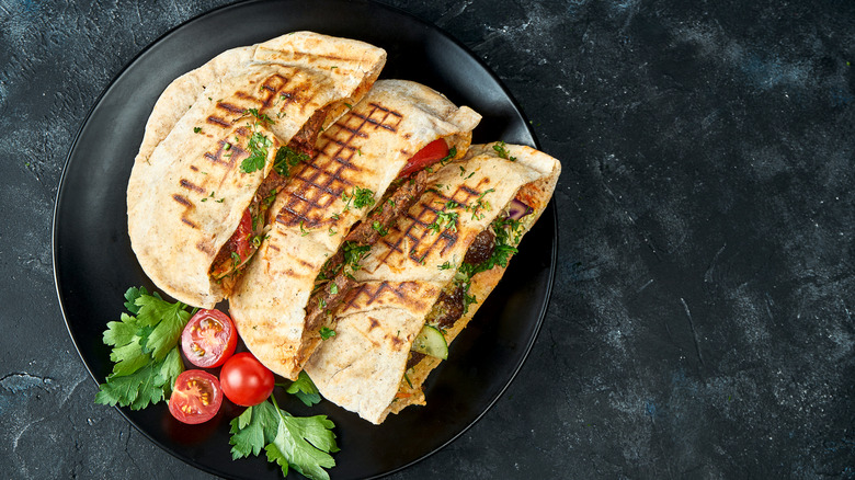 three flat bread sandwiches on a black plate with a sliced cherry tomato and parsley garnish