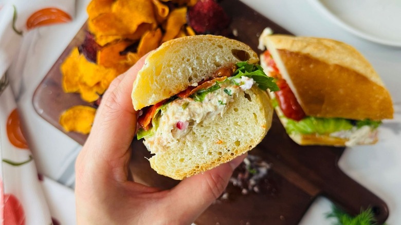 Hand holding half of an elevated tuna salad sandwich with chips and sandwich in the background