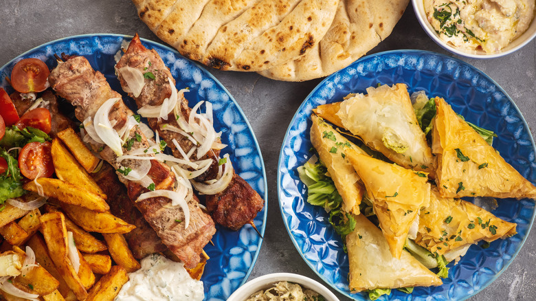 A table spread of Greek food with pita, hummus, skewers, and spanakopita