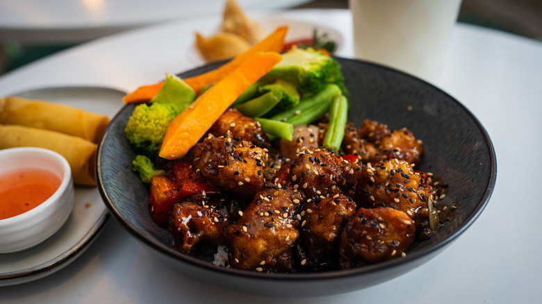 Fried chicken tossed in teriyaki sauce with sesame seeds with a side of green vegetables in a black bowl