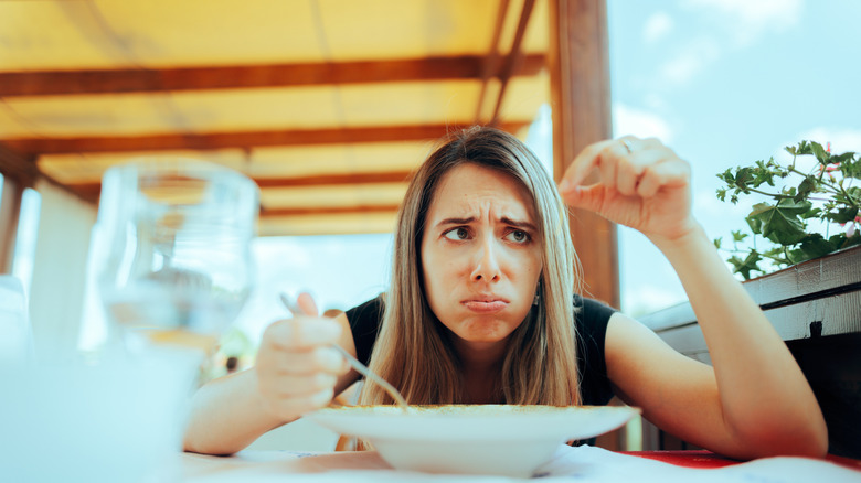 Woman seated at a table, frowning and looking at a piece of hair she's holding