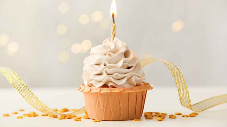 Birthday cupcake with burning candle on light table against blurred lights.