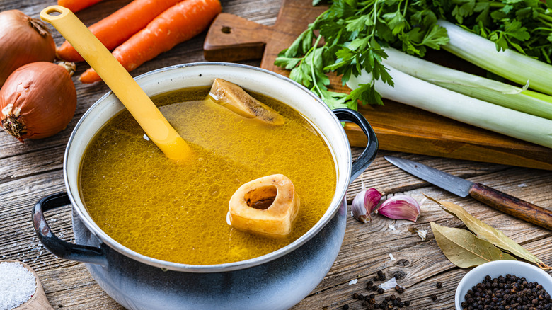 A high angle view of a pan of bone broth with a bone and ladle in it, surrounded by fresh vegetables
