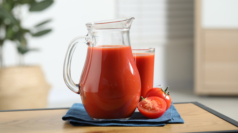 A pitcher and glass of tomato juice on a cloth next to a fresh tomato