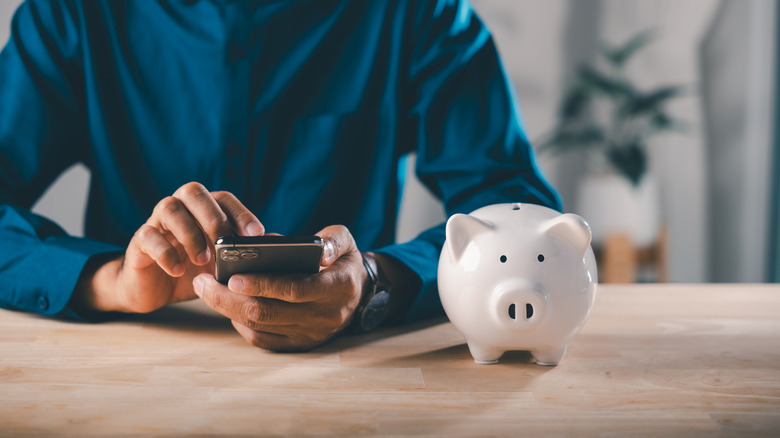 Person searching on cell phone with piggy bank on wooden table