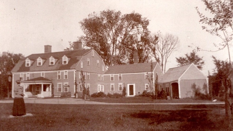 An old sepia-toned 1900s photo of The Wayside Inn with a woman standing in the foreground