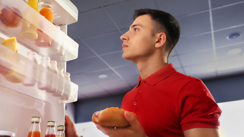 man holding hot dog looking into fridge