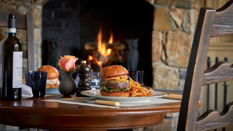A burger on a table with a bottle of wine. A roaring open hearth in the background