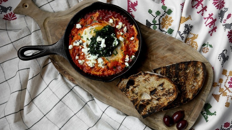 A pan of shakshouka on a wooden cutting board with bread and olives