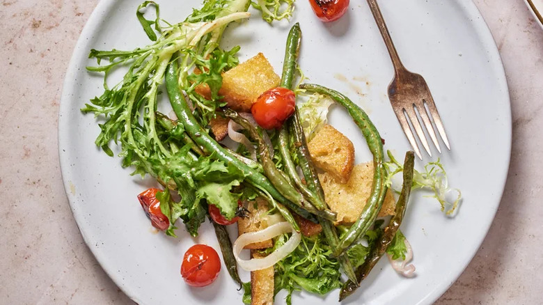 A white plate of green bean and tomato panzanella salad with a fork on the side