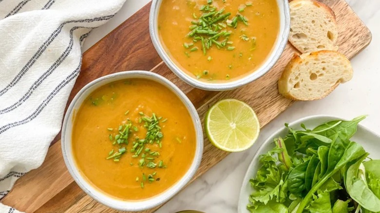 Two white bowls of roasted butternut squash soup with fresh parsley, half a lime, and slices of bread on a wooden cutting board