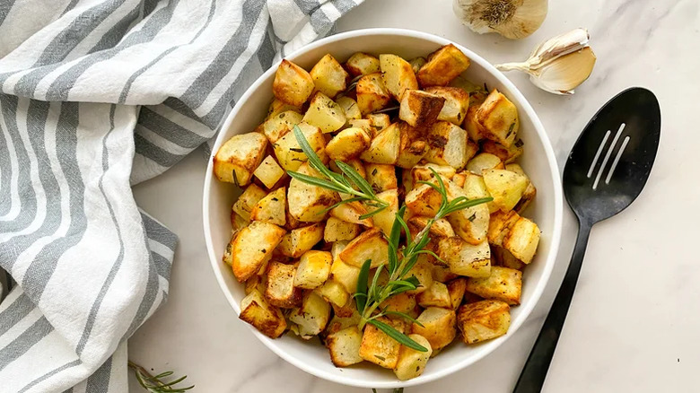 A white bowl of rosemary and garlic roasted potatoes with a black serving spoon next to it