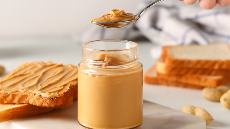 Spoonful of peanut butter held above a glass jar of peanut butter with bread in background.