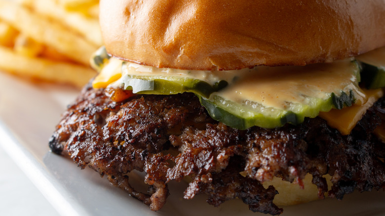 A closeup view of a crispy smashburger with pickles; a pile of fries is blurred in the background.